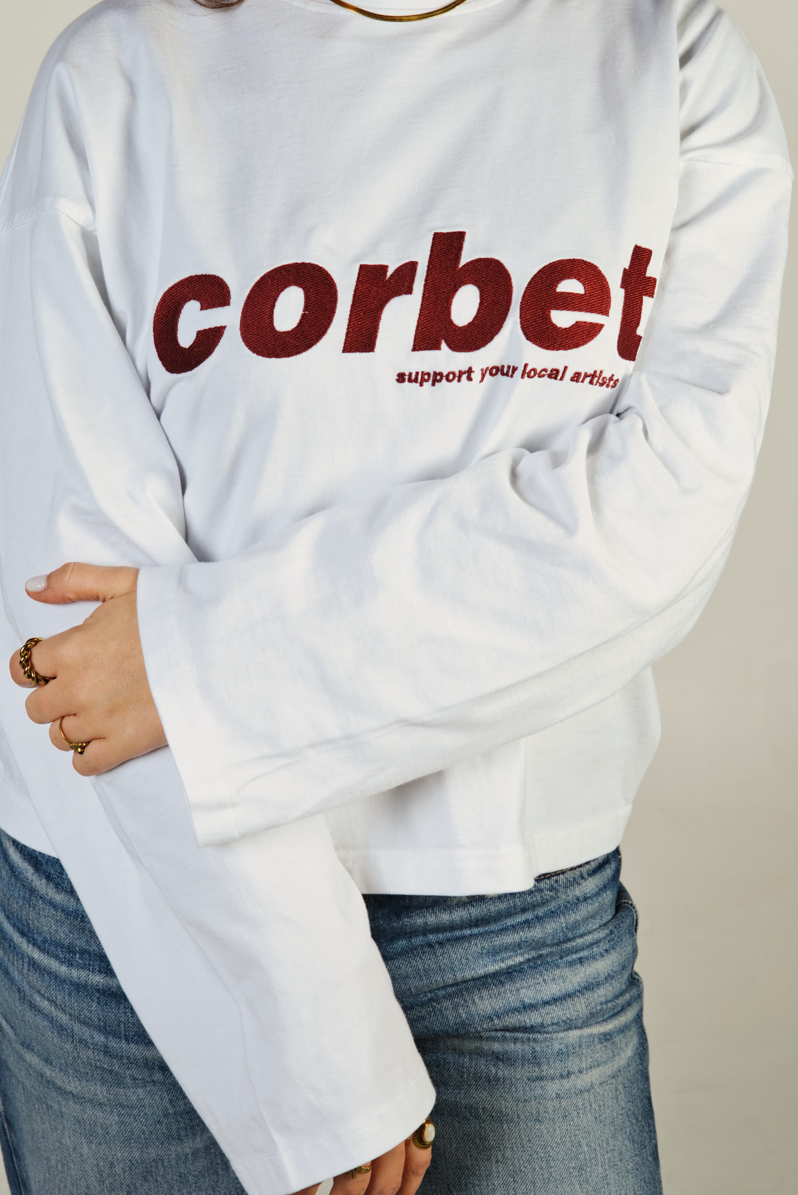 Person wearing a white long sleeve with 'corbet' embroidered in red on a plain background