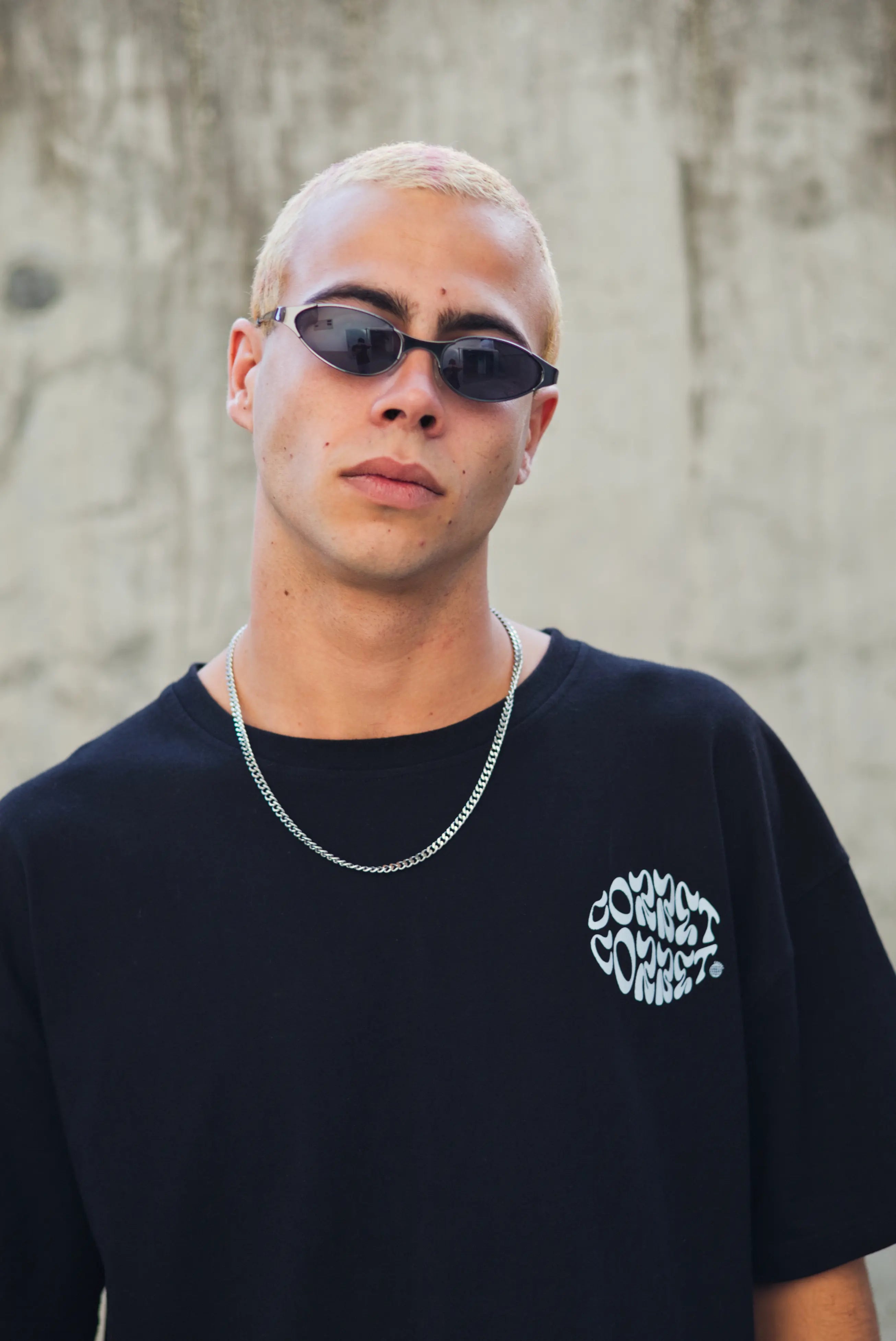 Person wearing sunglasses and a black t-shirt with text, standing against a textured wall.