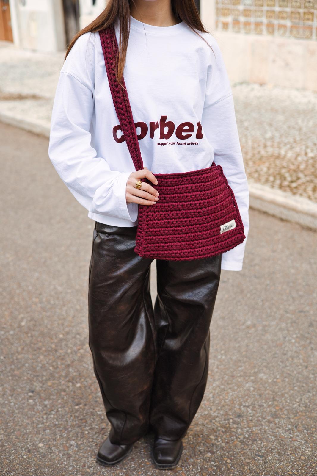 Isabel Aires wearing a white long sleeve with a red wine Corbet logo, brown pants, and carrying a matching red wine crochet bag.