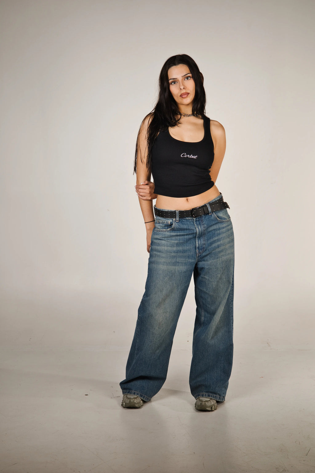 Woman wearing a black tank top with 'Corbet' text and blue jeans on a plain background