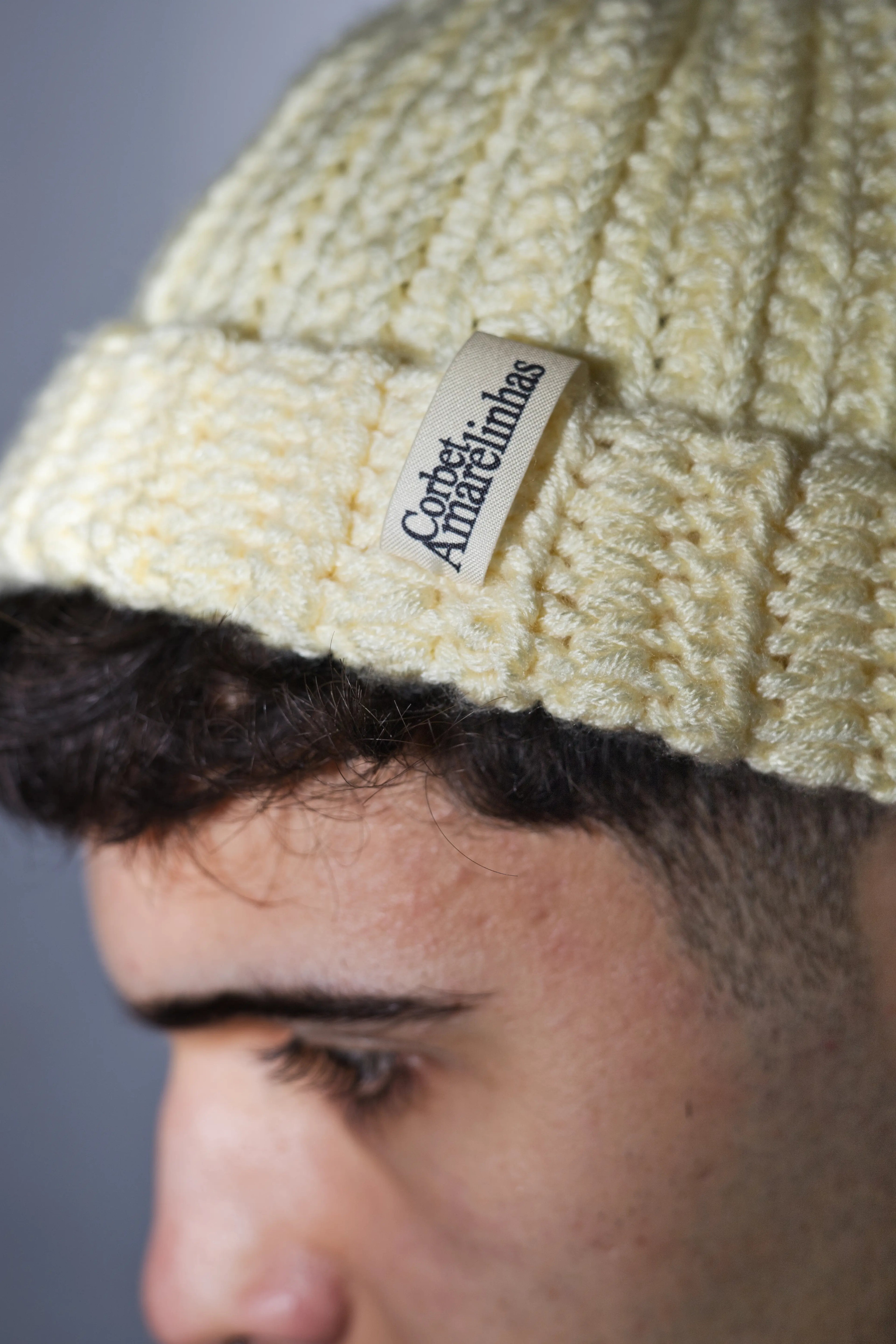Close-up of a person wearing a crochet beige beanie with a "Corbet x Amarelinhas" tag.