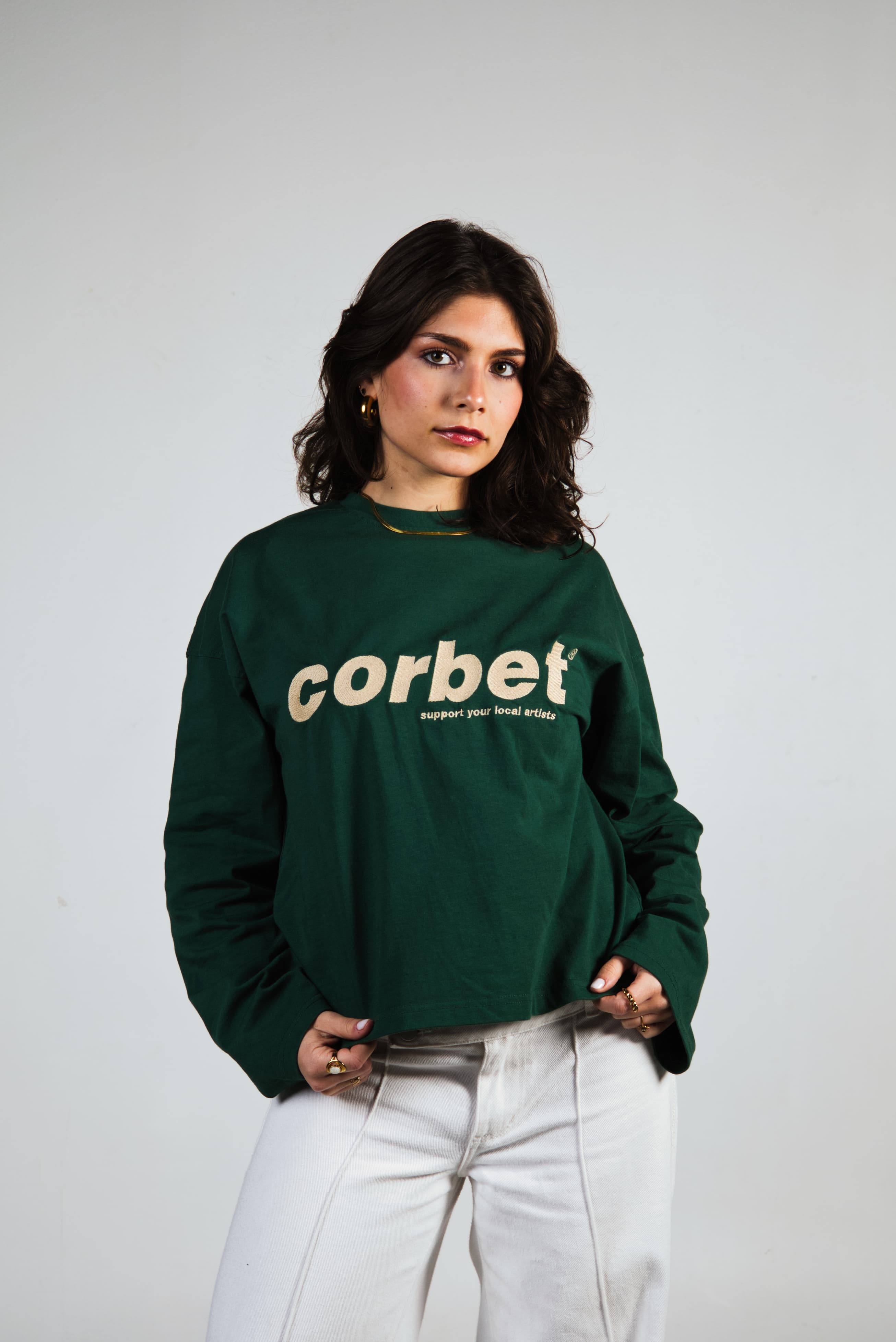 Woman wearing a green longsleeve with 'corbet' embroidered logo on a plain background