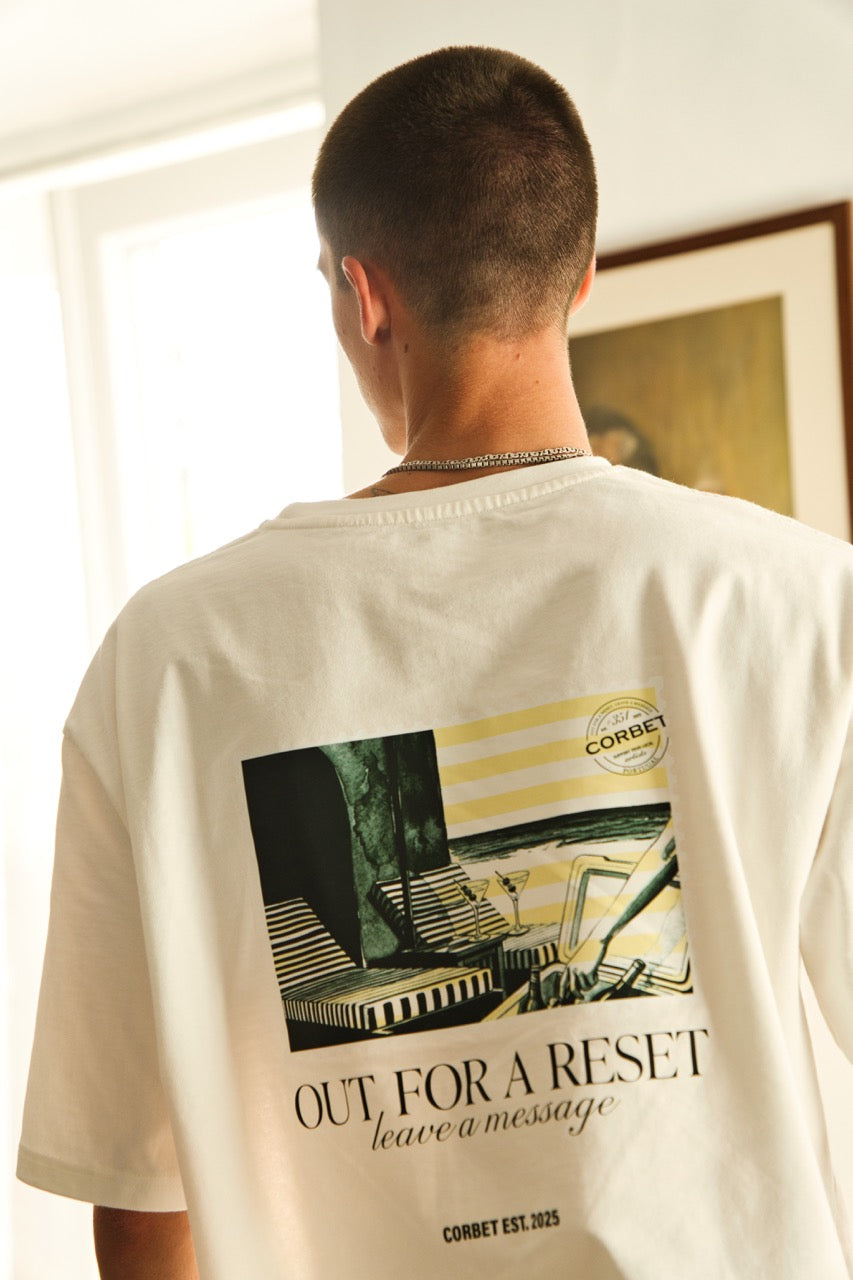 Person wearing a white t-shirt with a graphic design of a postcard and text on the back "Out for a reset, leave a message"