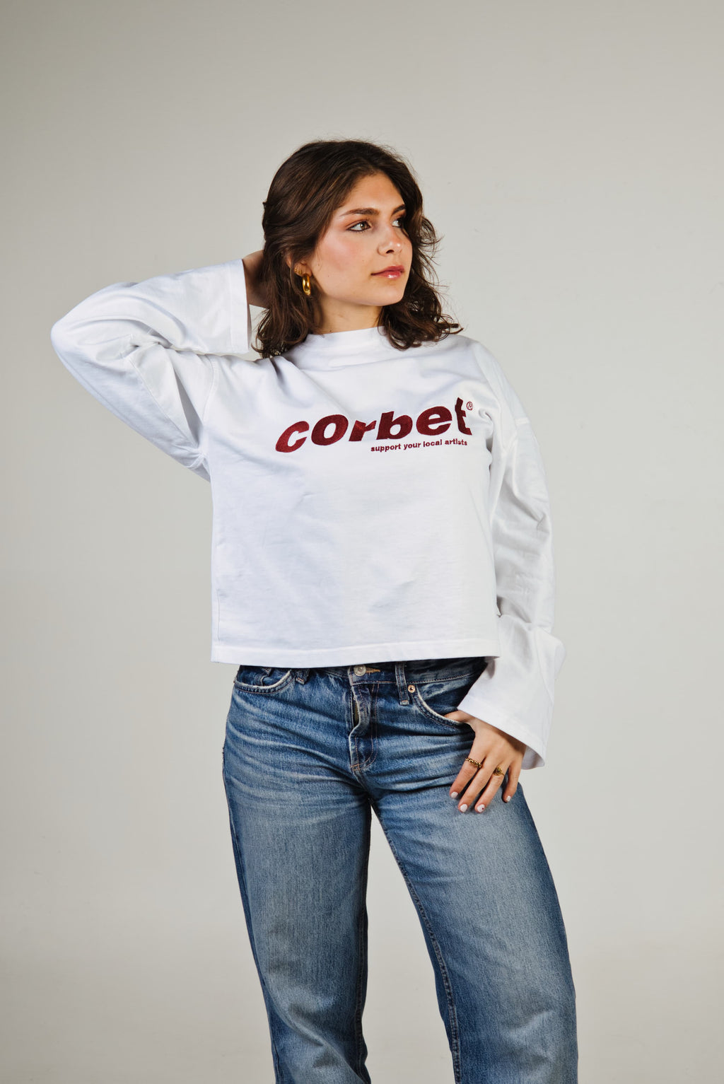 Person wearing a white long sleeve with 'Corbet' text and blue jeans on a plain background
