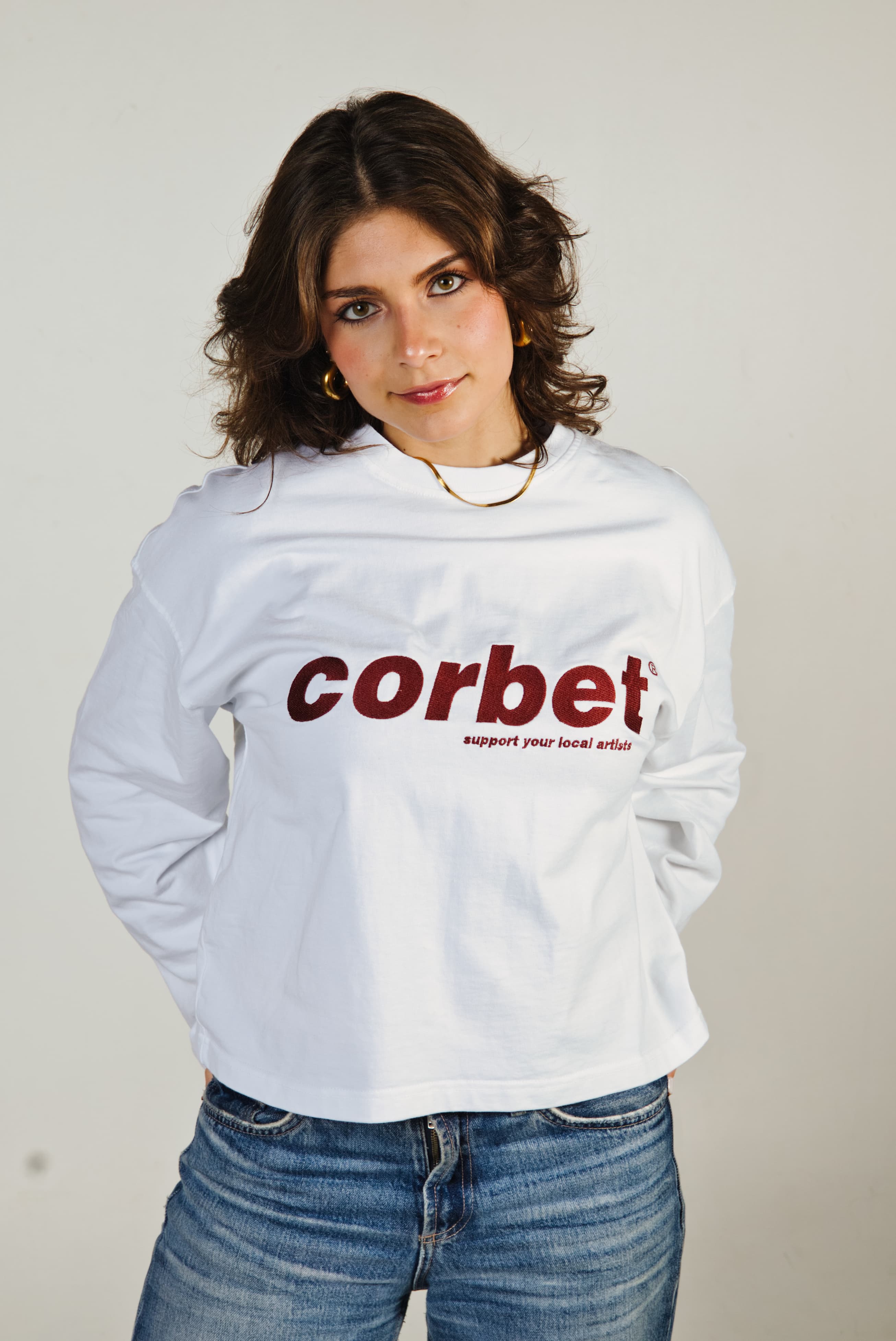 Person wearing a white long-sleeve with 'corbet' printed on it against a plain background
