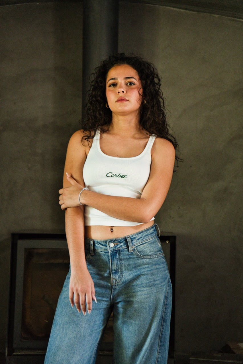 Woman wearing a white tank top and blue jeans standing against a dark background